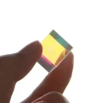 Piccolo cubo di vetro ottico colorato, trasparente con riflessi iridescenti, tenuto tra le dita. Perfetto come decorazione scientifica o oggetto da collezione.