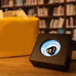 Oggetto scientifico in scatola con illustrazione digitale, posizionato su un tavolo in un ambiente con scaffali pieni di libri sullo sfondo.