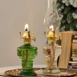 Due lampade di olio vintage con fiamma accesa, realizzate in vetro verde e trasparente, posizionate su un piatto di metallo. Perfette come decorazioni scientifiche o oggetti da collezione.