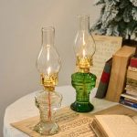 Due lanterne a olio vintage con vetro trasparente e base in vetro verde, posizionate su un tavolo con libri e decorazioni natalizie sullo sfondo.