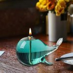 Decorazione in vetro a forma di narvalo con candela accesa, perfetta come oggetto decorativo scientifico o da collezione, su superficie in legno con fiori in sfondo.