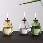 Tre vasi decorativi con candele scientifiche, in vetro colorato (giallo, trasparente, verde), con fiamme accese, su superficie nera, con foglia di pianta verde in sfondo.