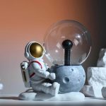 Modello di astronauta in miniatura accanto a una lampadina decorativa, simbolo di esplorazione e innovazione scientifica. Perfetto come decorazione per appassionati di scienza e spazio.