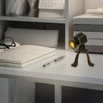 Microscopio da tavolo in miniatura con luce accesa, posizionato su una scrivania con libri e oggetti scientifici sullo sfondo. Perfetto come decorazione scientifica o oggetto da collezione.