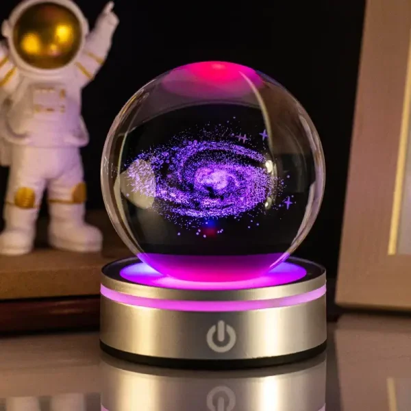 Palla di cristallo con rappresentazione di una galassia luminosa e colorata, posizionata su base con pulsante di accensione. Decorazione scientifica affascinante e moderna.