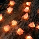 Catena di luci decorative a forma di pietre di sale rosa, posizionate su un tronco di legno naturale, creando un'atmosfera calda e accogliente. Perfette per decorazioni di ambienti interni o esterni.