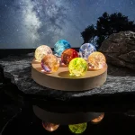 Palline luminose in vetro colorato su base di legno, ambientazione notturna con cielo stellato e natura, decorazioni scientifiche e oggetti luminosi per ambienti interni ed esterni.