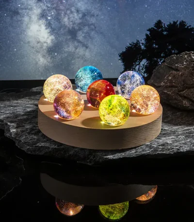 Palline luminose in vetro colorato su base di legno, ambientazione notturna con cielo stellato e natura, decorazioni scientifiche e oggetti luminosi per ambienti interni ed esterni.