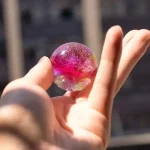 Palla di vetro decorativa con glitter rosa e dorato, tenuta tra le dita di una mano. Sfondo sfocato di un ambiente urbano.