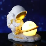 Figurina di astronauta in stile cartone animato che tiene un pianeta luminoso con anello, decorazione scientifica per ambienti interni.