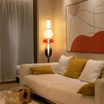 Lampada decorativa moderna con design elegante e illuminazione calda, posizionata accanto a un divano beige con cuscini color senape, in un soggiorno accogliente e di stile contemporaneo.