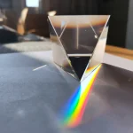 Piramide di cristallo trasparente che proietta un arcobaleno di luce sul piano sottostante, creando effetti ottici e giochi di riflessi.