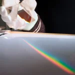 Immagine di un esperimento scientifico con un fascio di luce che crea un arcobaleno su una superficie lucida, con una mano che tiene un prisma o un oggetto ottico.