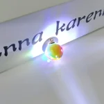 Pendente decorativo con effetto arcobaleno, illuminato da luci colorate, posizionato sotto una scritta personalizzata. Perfetto come decorazione scientifica o regalo originale.