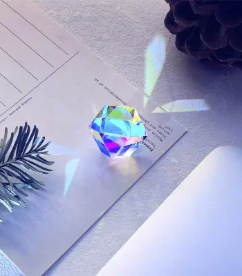 Cristallo decorativo di oggetto scientifico con riflessi iridescenti, posizionato su carta bianca con foglie e pigne intorno, ideale come decorazione da collezione o da scrivania.