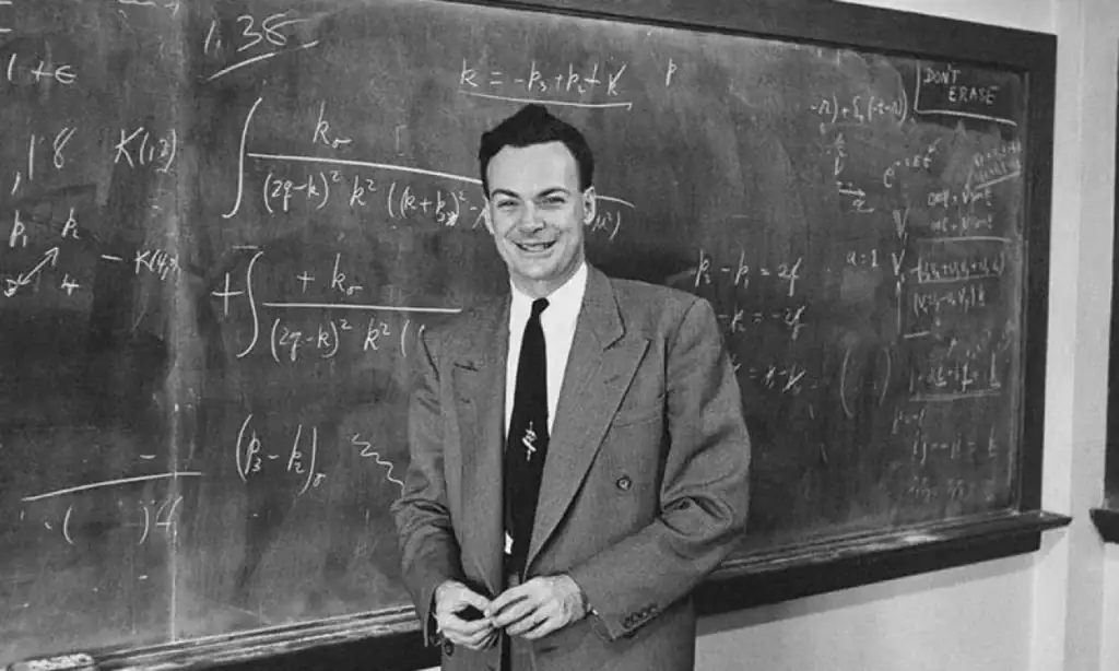 Uomo sorridente in abito davanti a lavagna con formule matematiche, oggetti scientifici decorativi e strumenti di laboratorio.