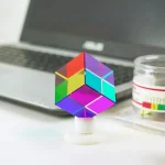 Cubo di Rubik con colori vivaci posizionato su una superficie con laptop e contenitore di oggetti scientifici sullo sfondo.