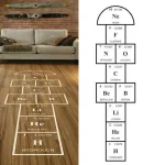 Tavolo periodico degli elementi con simboli e numeri atomici, rappresentato come un disegno su un pavimento in legno, con sfondo di divano e parete in mattoni.