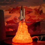 Razzo in miniatura con effetto fiamma arancione, simbolo di esplorazione spaziale e tecnologia, posizionato su una base nera con sfondo di nuvole rosse e arancioni al tramonto.