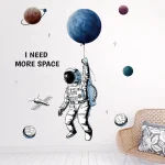 Murale decorativo con astronauta che tiene un pallone a forma di pianeta, circondato da pianeti e stelle, con la scritta "I NEED MORE SPACE" in stile grafico moderno.