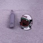 Punto 2: Penna USB grigia e spille con design di un casco spaziale sovietico e un volto di astronauta, perfetti come gadget scientifici e decorazioni a tema spaziale.
