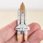 Spilla a forma di razzo spaziale in metallo con dettagli colorati, perfetta come decorazione scientifica o accessorio per appassionati di spazio e tecnologia.