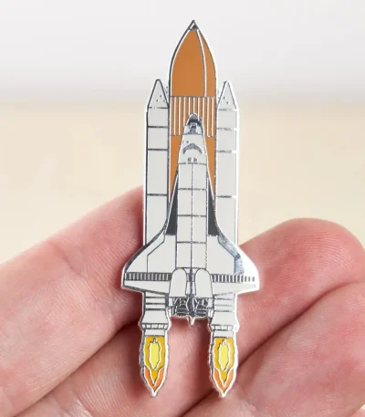 Spilla a forma di razzo spaziale in metallo con dettagli colorati, perfetta come decorazione scientifica o accessorio per appassionati di spazio e tecnologia.