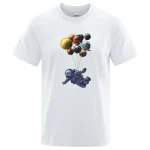 T-shirt bianca con stampa di un astronauta che vola tra pianeti e sfere planetarie colorate, ideale per appassionati di spazio e scienza.