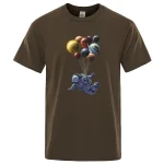 T-shirt color marrone con stampa di un astronauta che vola tra pianeti e sfere planetarie colorate, perfetta per gli appassionati di spazio e scienza.