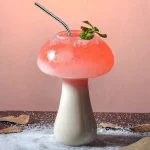 Bicchiere a forma di fungo con cocktail rosa, decorato con foglie di menta e cannuccia, su sfondo rosa pastello.