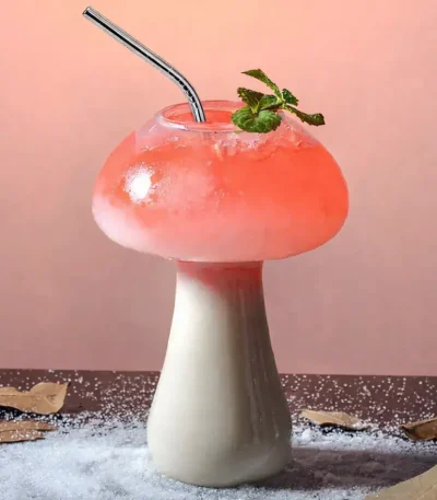 Bicchiere a forma di fungo con cocktail rosa, decorato con foglie di menta e cannuccia, su sfondo rosa pastello.