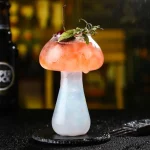 Cocktail decorativo a forma di fungo scientifico, con base bianca e cappello rosa, decorato con foglie e ciliegina, ideale come oggetto decorativo o per eventi tematici scientifici.