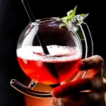 Cocktail in bicchiere di vetro trasparente con decorazione di foglie verdi e cannuccia nera, sfondo scuro.