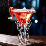 Cocktail colorato con decorazione a forma di medusa, servito in un bicchiere scientifico con tentacoli di vetro trasparente, su un piano di legno.