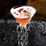 Cocktail rosa in bicchiere di vetro con decorazioni scientifiche, su superficie nera con riflessi lucidi.