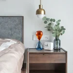 Vaso decorativo in vetro con sabbia arancione e blu, accompagnato da pianta di eucalipto in vaso, su comodino moderno in camera da letto.
