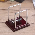 Pendo di Newton, modello scientifico in miniatura con sfere metalliche e supporto in legno, rappresenta il principio di conservazione dell'energia e il movimento delle sfere.