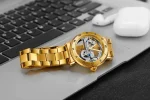 Orologio da polso con cassa dorata e movimento scheletrato visibile, posizionato su un laptop con tastiera nera e auricolari wireless bianchi sullo sfondo.