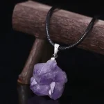 Collana con pendente in cristallo di ametista naturale, pietra viola con forma grezza, su cordino nero, su sfondo scuro.