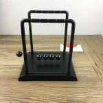 Péndolo di Newton in miniatura con cinque sfere nere appese, posizionato su una superficie di legno chiaro, ideale come decorazione scientifica o oggetto educativo.