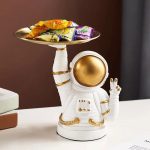 Decorativo astronauta in ceramica bianca con dettagli dorati, che tiene un piatto con snack colorati, ideale come oggetto decorativo scientifico e originale.