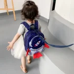 Bambina con zaino a tema spaziale, cammina in un ambiente interno, vista da dietro, con capelli ricci e abbigliamento casual.