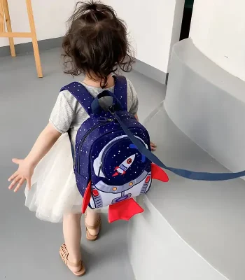 Bambina con zaino a tema spaziale, cammina in un ambiente interno, vista da dietro, con capelli ricci e abbigliamento casual.