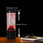 Vaso di laboratorio con bolle rosso vivo, altezza 25,5 cm e diametro 9 cm, posizionato su un tavolo di legno con occhiali e quaderno accanto, sfondo nero.