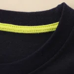 Primo piano di una maglietta nera con cucitura interna fluorescente giallo lime, evidenziando i dettagli di design e materiali.