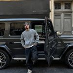 Giovane uomo in abbigliamento casual con cappello, posa davanti a un'auto nera in un contesto urbano, rappresentando uno stile moderno e tecnologico.